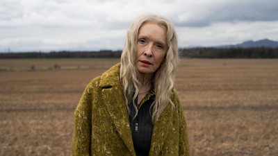 Photo of Lindsay Duncan in character as Ollie. Wearing a green wool coat, she stands in a field.