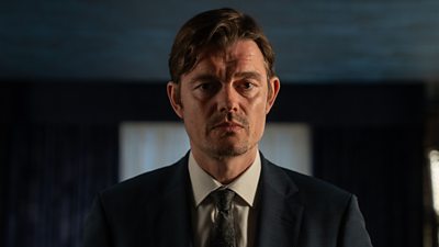 Photo of Sam Riley in character as Dylan. Wearing a suit, he stands in a darkened room. 