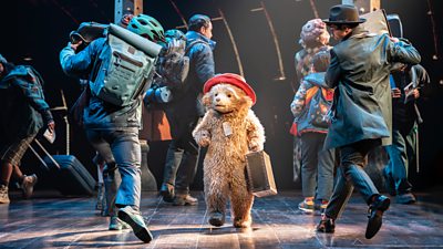 A person in a bear suit stands on stage holding a suitcase as other performers carrying backpacks and suitcases mill around them