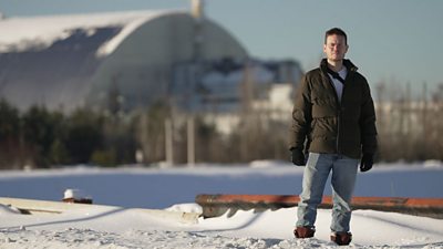 A man, BBC journalist Jordan Dunbar, stands on snowy ground with Chernobyl visible in the background behind him