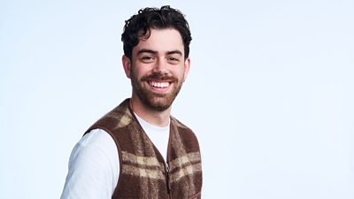A man in a brown zip up waistcoat and white top smiles to camera