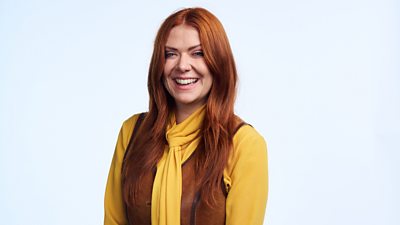 A woman with long red hair smiles to camera, wearing a brown waistcoat over a yellow top