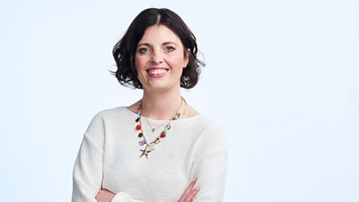 A woman whit short brown hair smiles to camera, wearing a white top and a colourful necklace.
