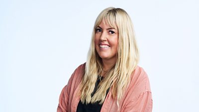 A woman with long blonde hair smiles to camera, wearing a pink shirt over a black top