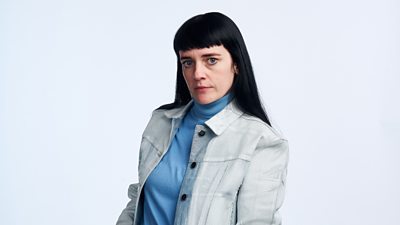 A woman with long black hair looks to camera, wearing a blue rollneck top beneath a light denim coloured shirt