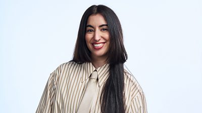 A woman with long brown hair smiles to camera wearing a pale shirt with brown stripes and a pale cream tie.