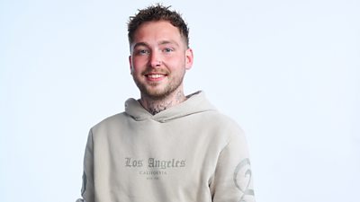A man with short brown hair smiles to camera wearing a cream/beige hoodie