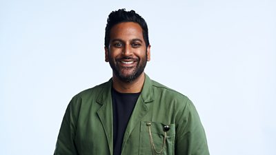 A man in a black t-shirt and green shit smiles to camera