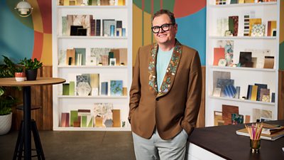 A man in a brown blazer, blue top and grey trousers stands in a brightly coloured design studio smiling