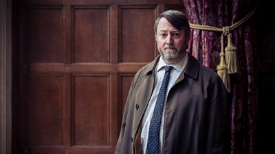 Photo of David Mitchell in character as John ‘Ludwig’ Taylor in a wood-panelled room.