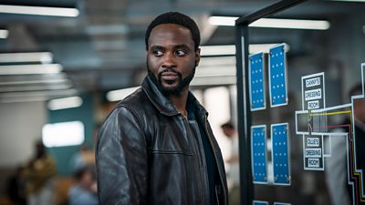 Photo of Dipo Ola in character as DI Russell Carter. He stands in an office beside a board with details of a police investigation.