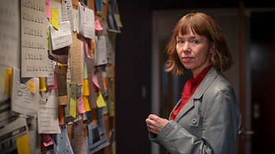 Photo of Anna Maxwell Martin in character as Lucy. She stands beside a corkboard full of paper documents.