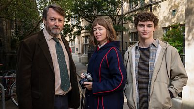 Photo of David Mitchell, Anna Maxwell Martin and Dylan Hughes in character as Ludwig, Lucy and Henry respectively. They stand outdoors. Lucy holds a camera.
