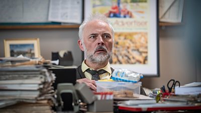 Mark Bonnar in character as Gareth Fisher. He sits behind a messy office desk, cluttered with stacks of papers, snacks, picture frames and stationary.