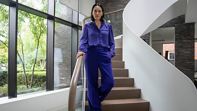 Sian Clifford in character as Joanne Kemper MP. Wearing a purple blouse and matching purple trousers, she walks down a white staircase in a modern home.