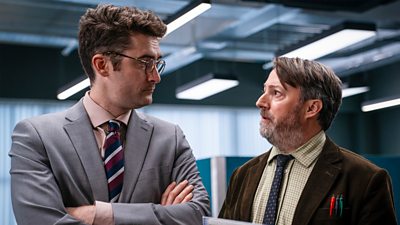 Ben Ashenden in character as DC Ethan Cole folds his arms and looks at David Mitchell in character as John ‘Ludwig’ Taylor.