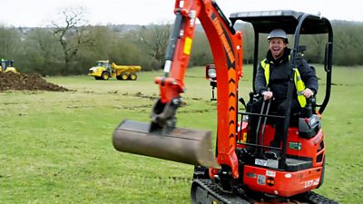 A man in a high vis jacket sits in a digger looking delighted
