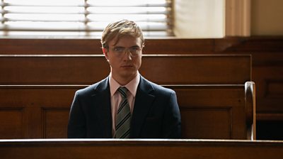 A blonde man waring glasses sits on a wooden bench in a suit