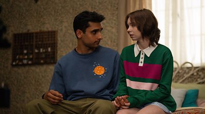 Zain Iqbal playing Ravi and Emma Myers playing Pip sitting on a bed holding hands 