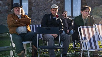 Four people sitting across rows of foldable chairs on grass outside a building. They all watch something offscreen.