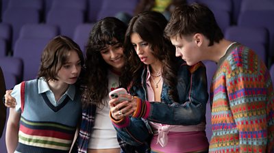 Pip, Cara, Lauren and Connor stand in a line and look at Lauren's phone. The four are standing it front of a row of purple seats