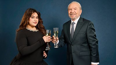 A woman in a black dress and a man in a dark suit smile to camera as they raise a toast with champagne flutes