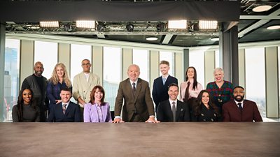 The Celebrity Apprentice candidates sit and stand either side of Lord Sugar at a boardroom table in a skyscraper