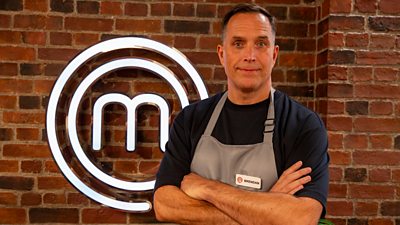 A man stands in the kitchen with his arms folded. He wears a grey apron over a dark top.