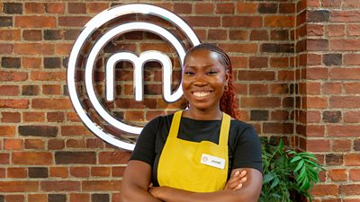 A woman in a dark t-shirt and yellow apron stands in the MasterChef kitchen smiling