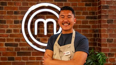 A man in a dark t-shirt and beige apron stands smiling in the kitchen with his arms folded
