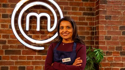A woman in a long sleeve marron top and a dark blue apron stands in the MasterChef kitchen smiling to camera