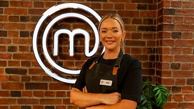 A woman in a dark t-shirt and black apron stands in the MasterChef kitchen smiling to camera