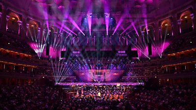 A photo from the last night of the BBC Proms 2023 - an orchestra can be seen surrounded by a crowd with bright purple lighting