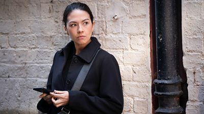 A woman in a dark coat stands against a wall with a phone in her hand. looking at something happening off screen