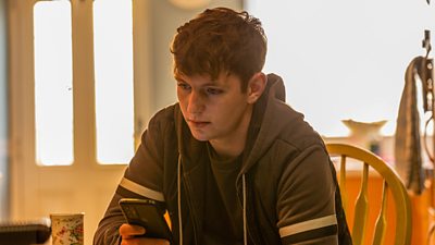 A young man sits at a kitchen table looking sad about something, holding a mobile phone