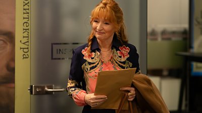 Lesley Manville as Susan Ryeland. She stands in an office and holds a paper envelope. 
