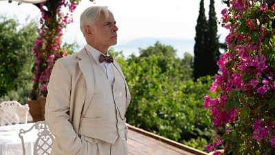 Mark Bonnar as Elmer Waysmith. Wearing a three-piece suit, he stands on a terrace above vibrant gardens. 