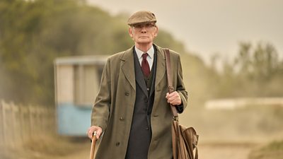 An older man wearing a dark coloured suite, a green overcoat and flatcap. He's walking down a dusty path