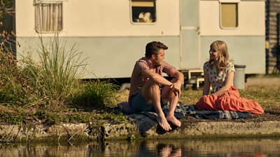 A mand and a woman sat on the side of a lake in the sun. Behind them is a white and blue faded caravan 