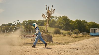 A young person dressed in a blue astronaut costume. They are walking along a dusty path next to a playground
