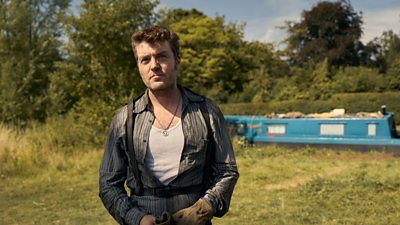 A man wwith dark hari is wearing a vest and a blue shirt. He is standing on some grass in front of a canal