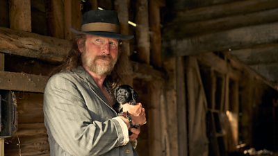 A man waeing an old jacker and hat stands in a barn holding a puppy