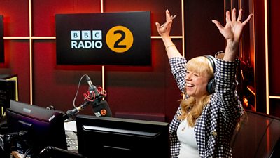 A woman in a check shirt sits in a radio studio, smiling with her arms raised in celebration