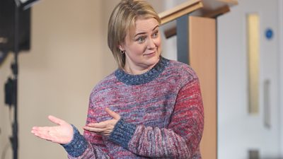 Philippa Dunne in character as Anne during filming of Amandaland. Wearing a colourful knitted jumper, she gestures to something offscreen. 