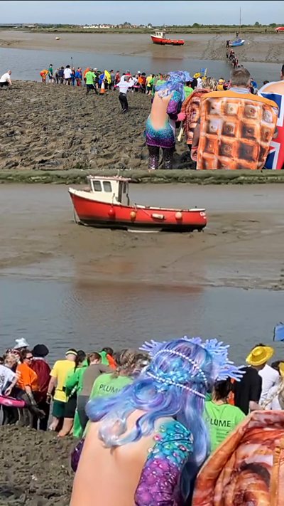 People in fancy dress walking through mud on a river bank in the sunshine