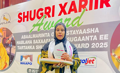 A woman journalist winning the Shukri Hariir Award Winner in Somaliland, she wears a navy headscarf and is holding a laptop and certificate - she is standing against a awards backdrop with bold colours of red and green in the text signage of the awards names, and sponsor/partner logos.