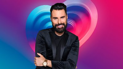 A photo of Rylan Clark in a black suit with a multi-coloured heart in the background 