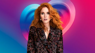 Angela Scanlon wearing a flower print jacket stood in front of a multi-coloured heart
