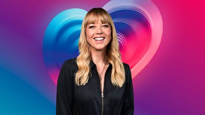 Sara Cox in a black jacket standing in front of a multi-coloured heart