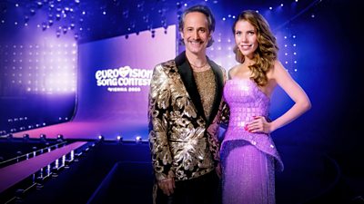 Eurovision host Victoria Swarovski wears a purple dress and host Michael Ostrowski wears a glittering back and gold suit. They are set against a depiction of the Eurovision Song Contest 2026 stage, bathed in purple light.